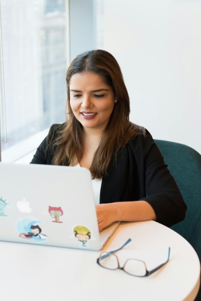 pexels-photo-1181292-1181292 Smiling woman in black blazer working on laptop with open notebook on desk in a modern office setting.