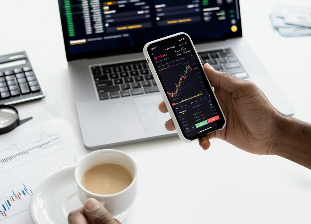 pexels-photo-6801874-6801874 A person trades stocks on a smartphone with a laptop, coffee cup, calculator, and documents in view.